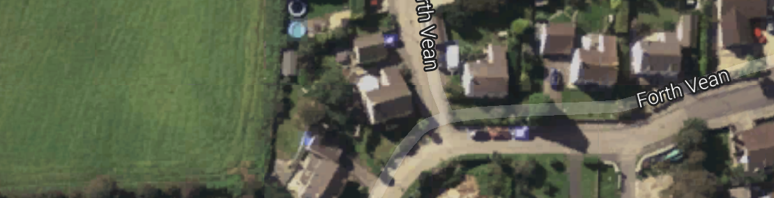 The image is a satellite view showing a T-shaped intersection on Forth Vean street with residential homes on either side. There is a mixture of greenery, including large fields, and various sized lawns. The houses are predominantly light brown, with some roofs appearing blue and gray. The image is clear, with well-defined boundaries between the houses, the street, and the grassy areas.